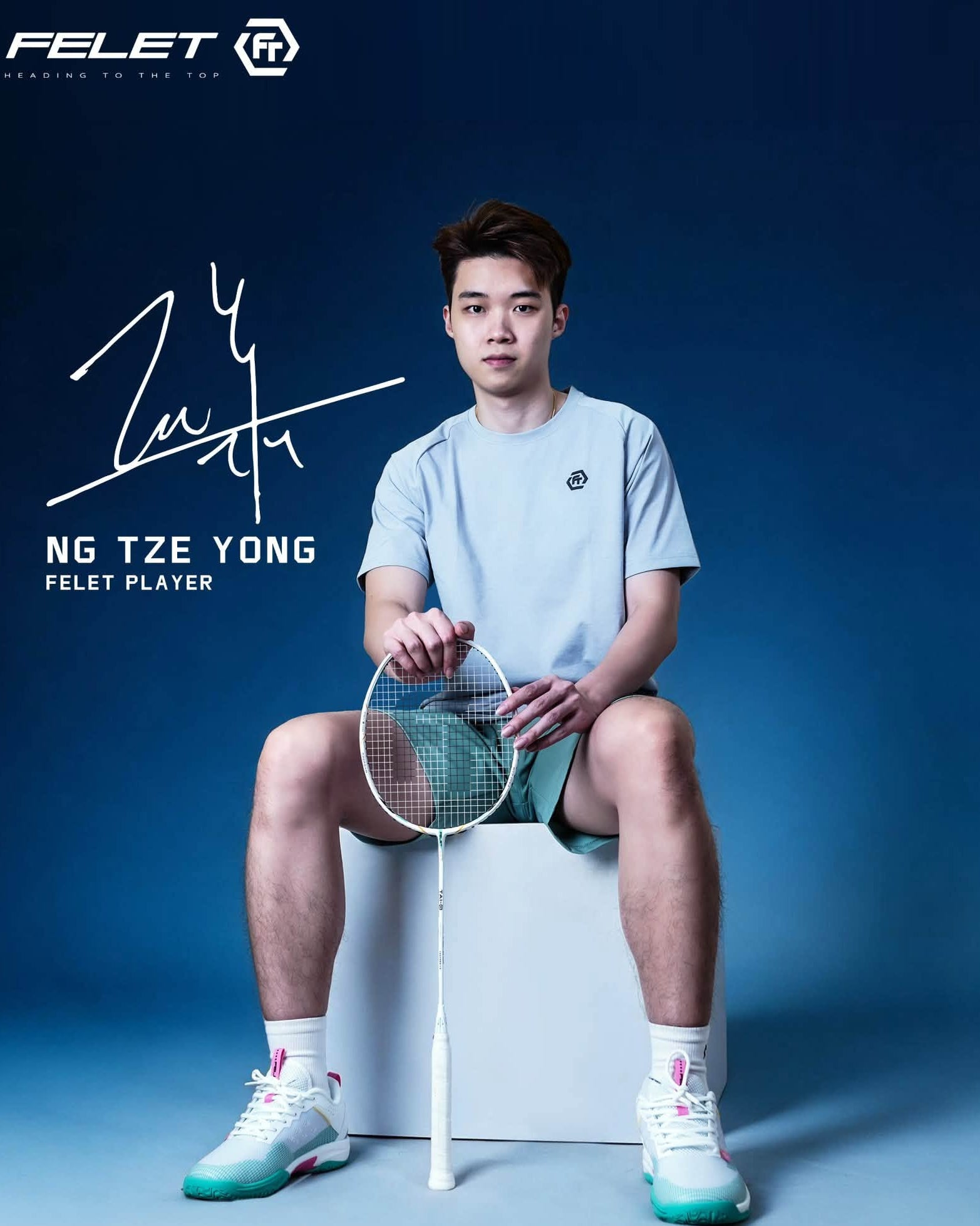 A badminton player Ng Tze 
Yong using Tai-G Aggressive White sits on a white block against a dark blue studio background, wearing a light blue short-sleeve athletic shirt, teal shorts, white socks, and white sneakers with teal accents. A badminton racket is held in front of him