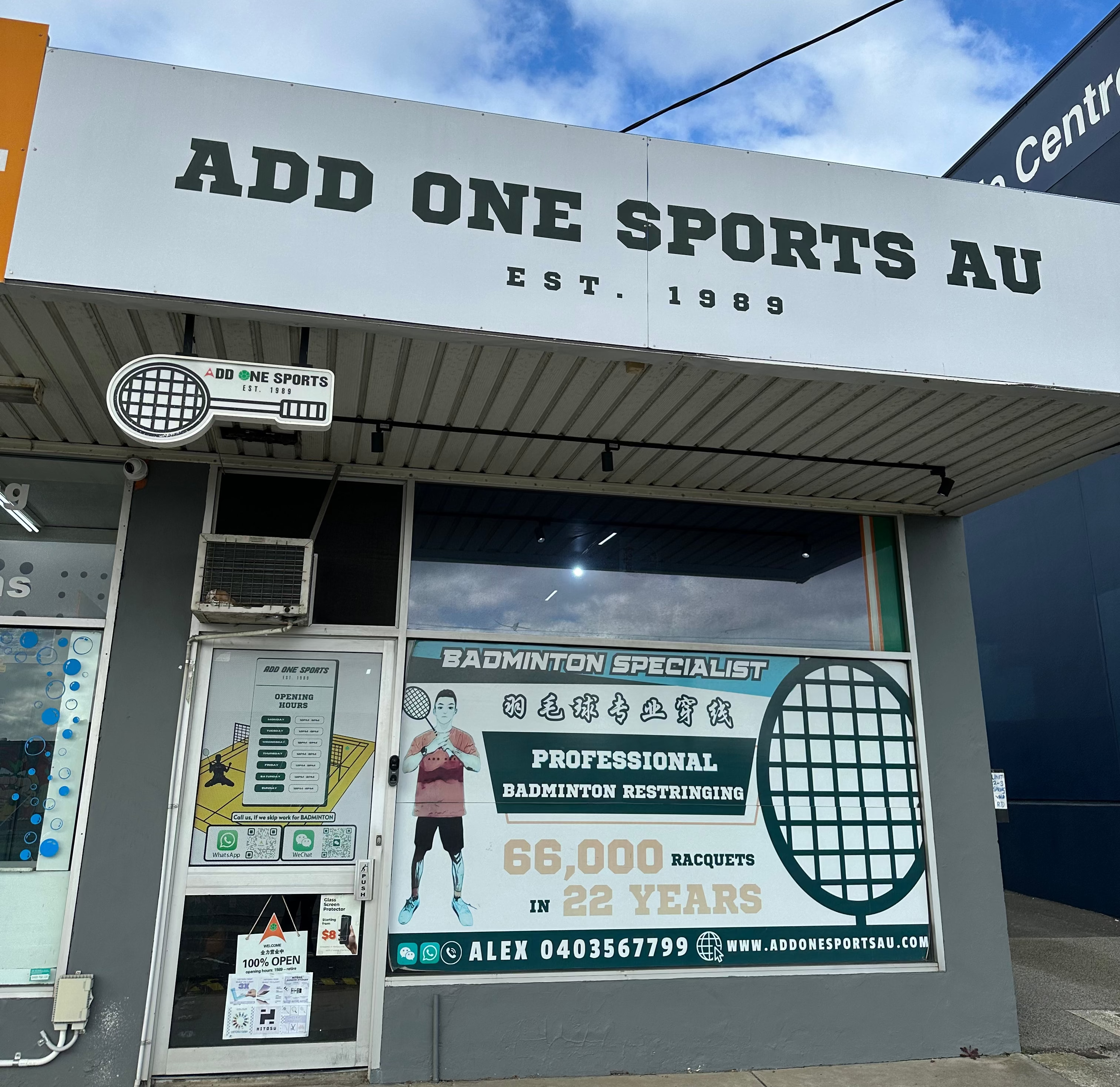 Front exterior of a sports shop named ADD ONE SPORTS AU, featuring a large white sign, glass entrance, and a prominent green and white badminton specialist banner on the storefront.